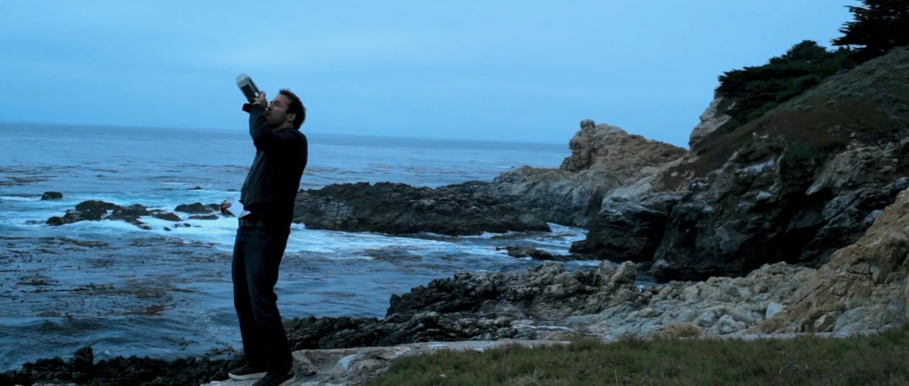 I Melt With You (2011) movie still: wide — A rocky coastal shoreline overlooks a dark ocean under an overcast blue-hour sky. A man stands on the…