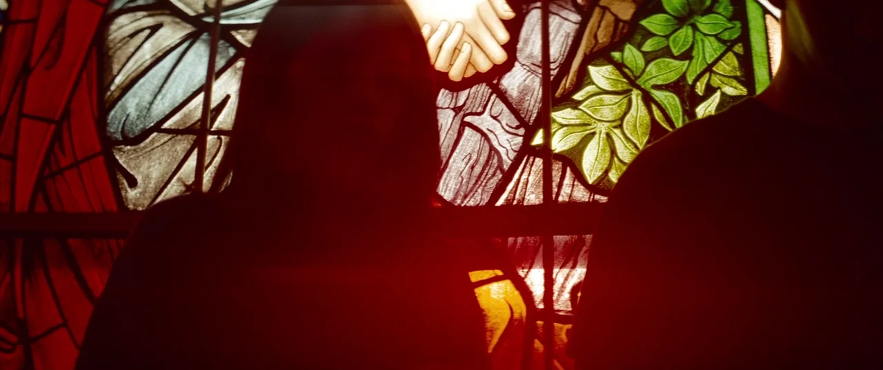 Spring Breakers (2012) movie still: medium — Two people stand in front of a colorful backlit stained glass window. The subjects are positioned in…