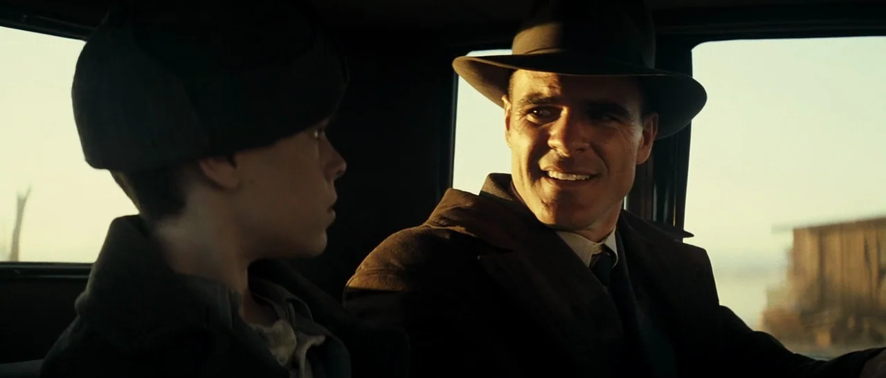 Changeling (2008) movie still: medium — The interior of a 1930s automobile with dark upholstery and window frames. A man wearing a fedora looks over…
