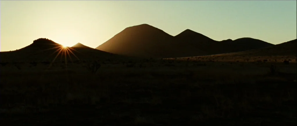 No Country For Old Men movie still: wide — A vast, arid desert landscape features a dark mountain range under a rising sun. The sun peeks over the…