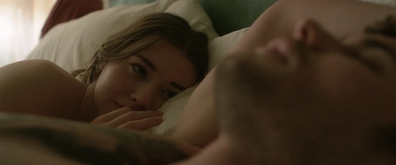 Marked Men: Rule + Shaw (2025) movie still: closeup — A light-filled room contains a bed with white pillows and neutral bedding. A woman rests her…