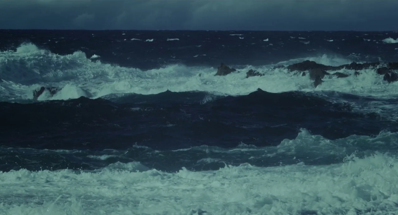 Julieta (2016) movie still: establishing — A turbulent ocean under a dark, overcast sky shows rough water crashing against rocks. Waves foam and…
