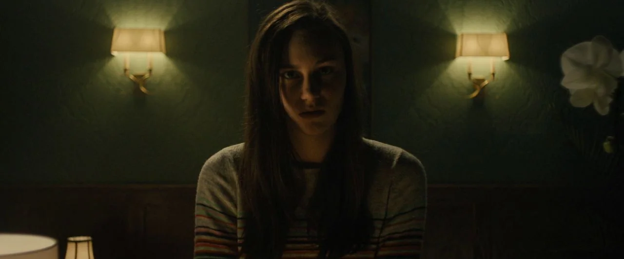 Freaky (2020) movie still: medium — A dark hotel room features dark green walls illuminated by two symmetrical wall sconces. A young woman with long…