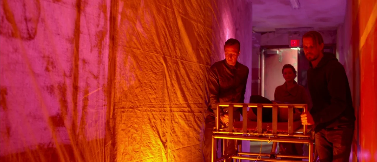 Rupture (2016) movie still: medium — A narrow hospital corridor is illuminated by vibrant red and orange artificial light sources. Two men and a…