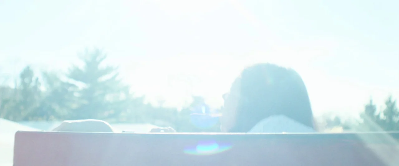 Wildling (2018) movie still: medium — A park bench is positioned outdoors against a background of bright sunlight and trees. A person sits on the…