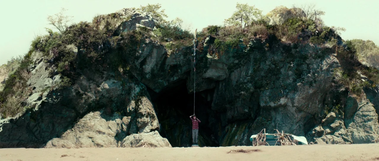 Swiss Army Man (2016) movie still: wide — A tall rocky cliff features a dark cave opening at the base on sand. A single person stands beneath the…