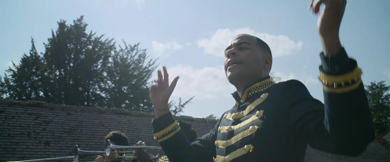 Cinderella (2015) movie still: medium — A man in a gold-trimmed uniform conducts music outdoors against a blue sky. Another musician plays a silver…