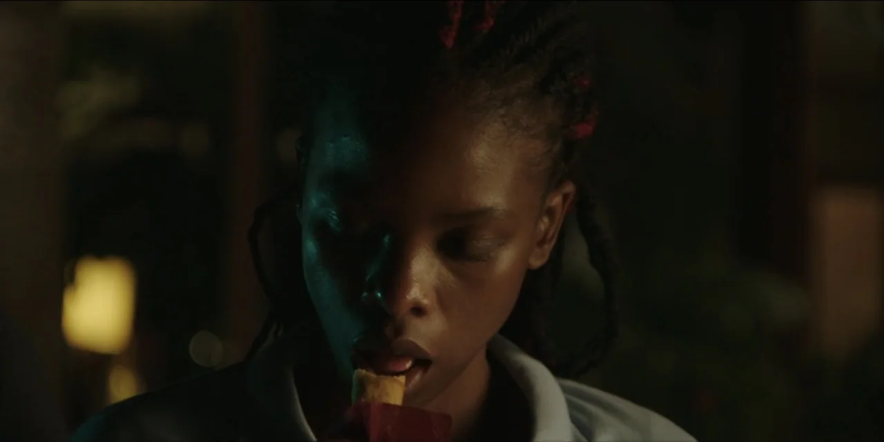 The Hunted (2024) movie still: closeup — A dimly lit room provides a dark background behind the subject. A young woman eats from a small red package…