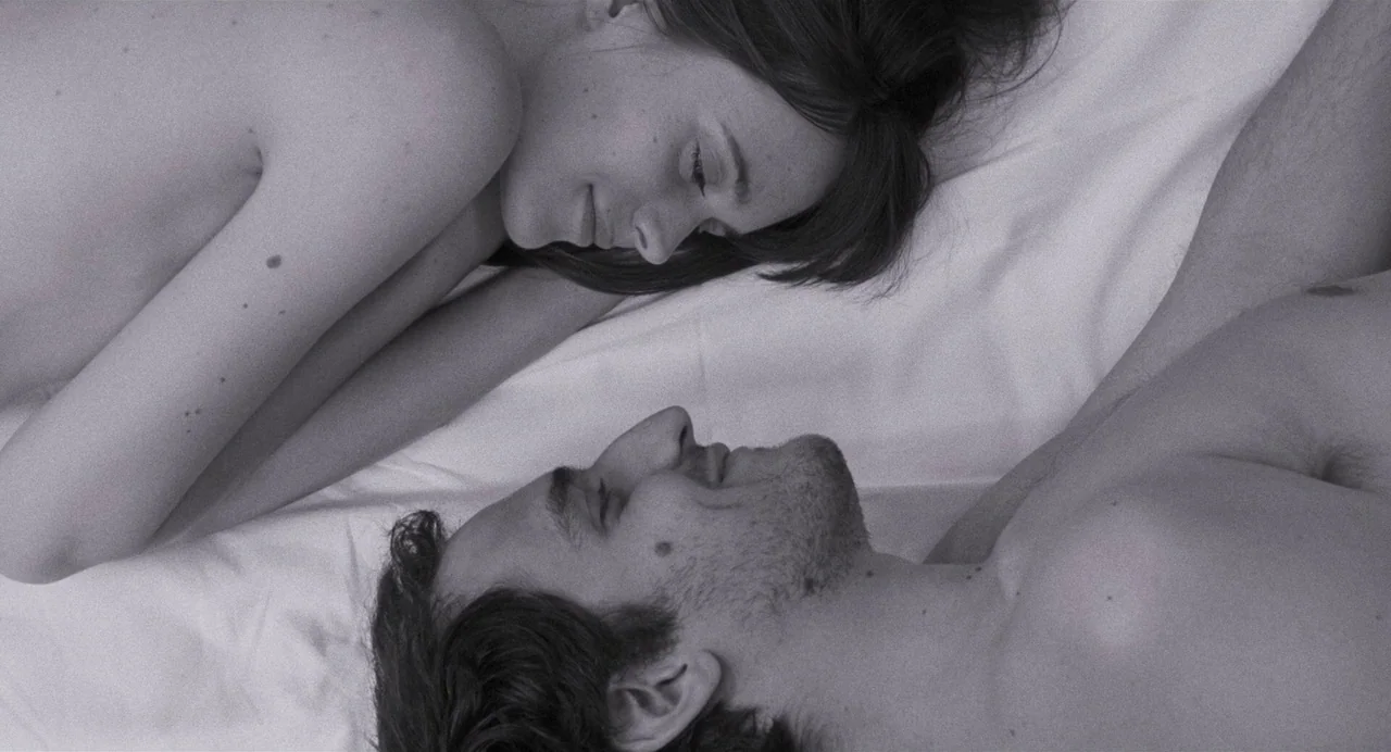 Godard Mon Amour (2015) movie still: medium — A man and woman lie face-to-face on a bed with white bedding. They are positioned in profile view…