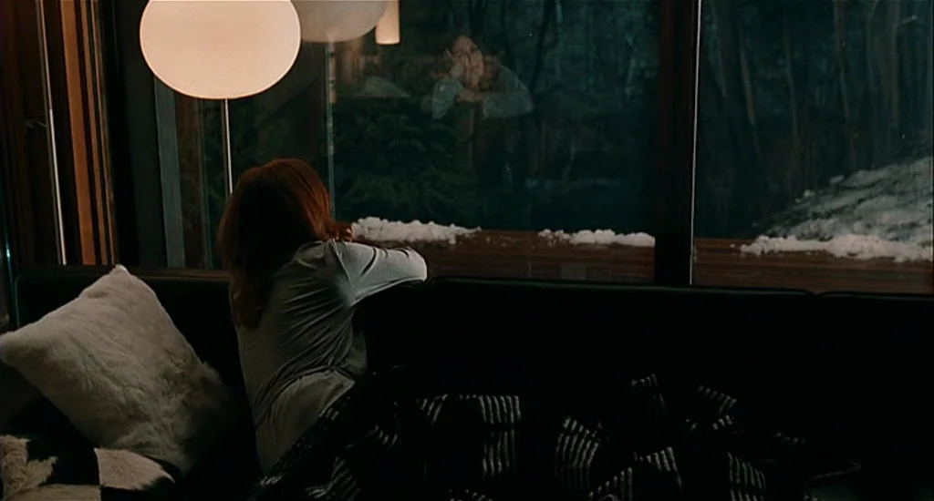 Chloe (2009) movie still: medium — A contemporary dark living room at night features a large glass wall looking outdoors. A woman sits on a sofa…