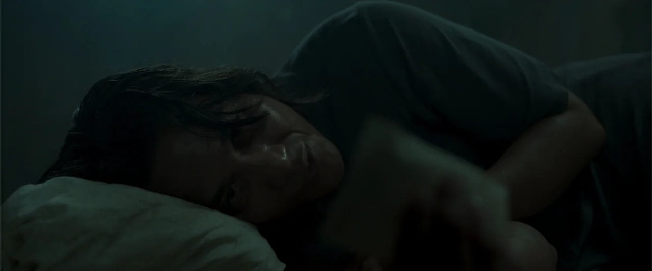 The Unholy (2022) movie still: closeup — A dimly lit room at night with a person lying on a pillow. The subject looks down at a small object while…