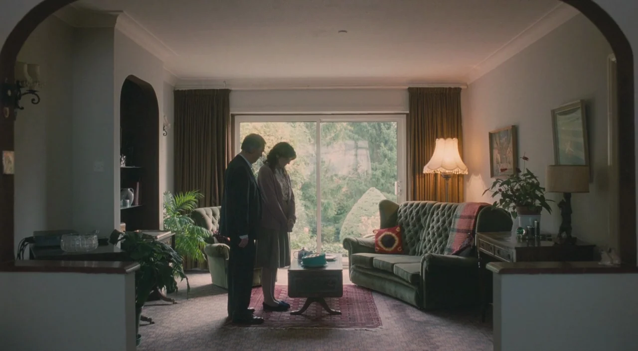 Broken (2012) movie still: wide — A 1970s interior with patterned carpet, heavy curtains, and a tufted green sofa. Two people stand facing each…