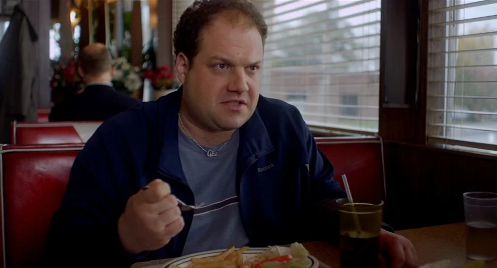 Dark Horse (2011) movie still: medium — A diner booth with red vinyl seating and light-colored wooden surfaces near a window. A man in a blue jacket…