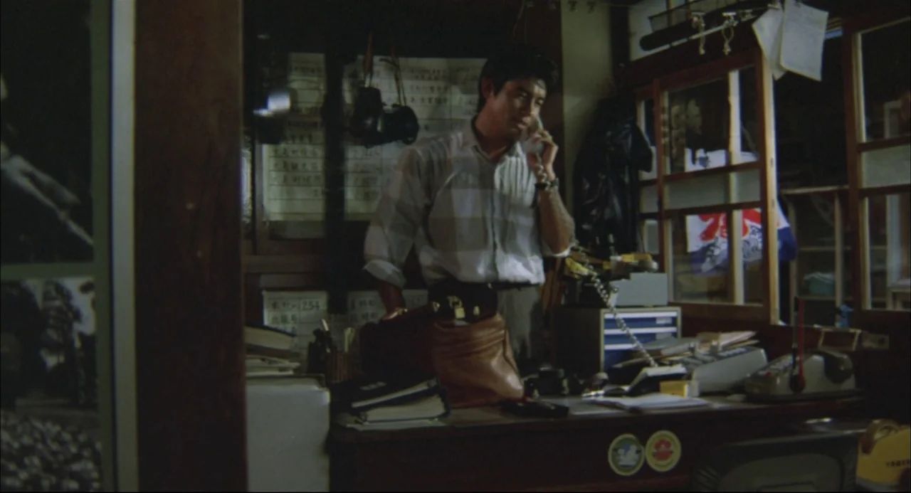 The Return of Godzilla (1984) movie still: medium — A cluttered small office space features a wooden desk piled with documents and equipment. A man…