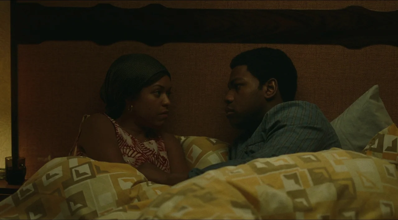 Red, White & Blue (2020) movie still: medium — A dimly lit bedroom features 1970s style wallpaper and patterned yellow bedding. A couple lies in the…