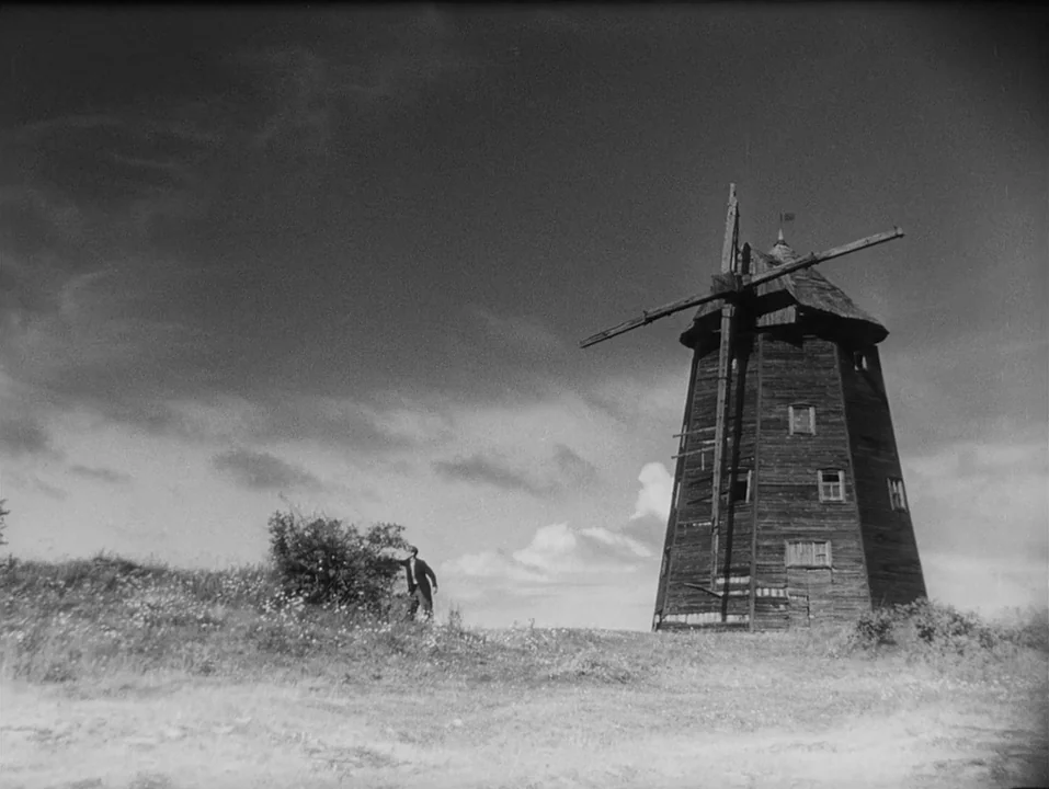 A Ship Bound for India (1947) movie still: wide — A rural landscape features a wooden windmill on a hill under a cloudy sky. A man in a dark suit…