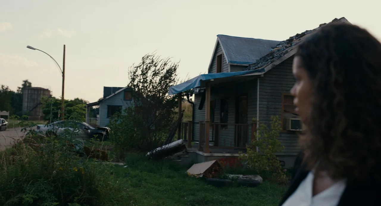 Barbarian (2022) movie still: medium — A desolate street features weathered wooden houses with blue tarps on their damaged roofs. A person stands in…