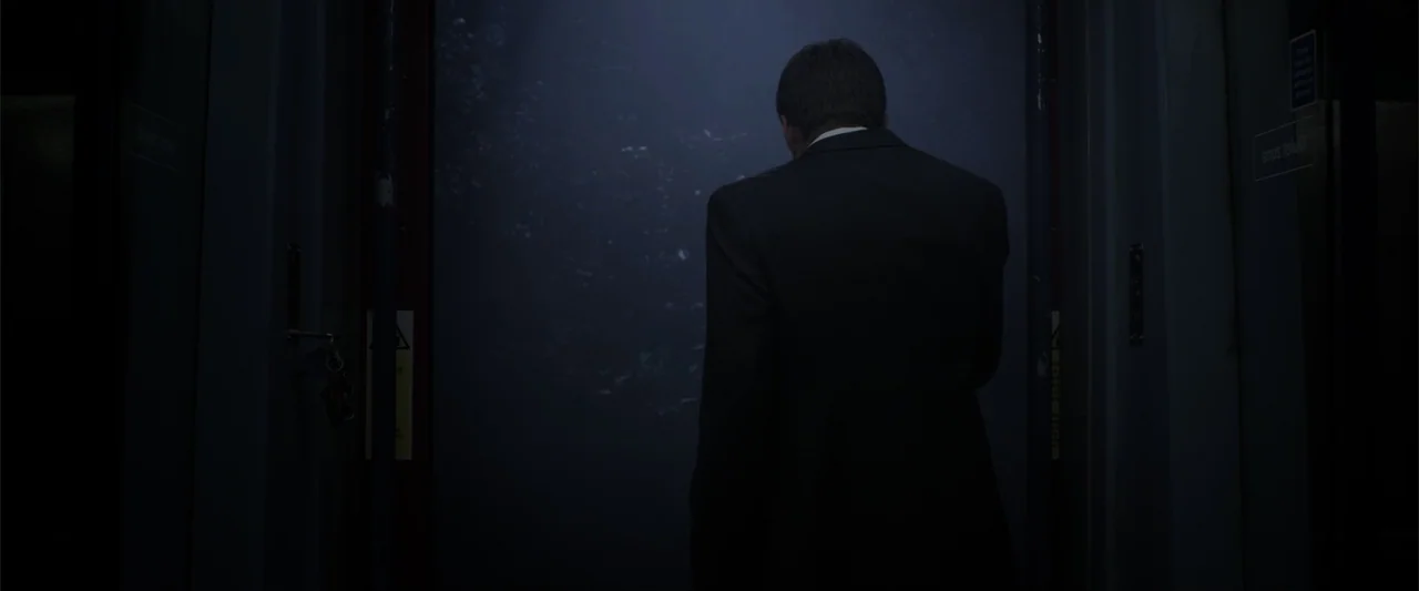 Howl (2015) movie still: medium — An elevator interior with dark metallic doors and a dimly lit control panel. A man in a suit stands facing the…