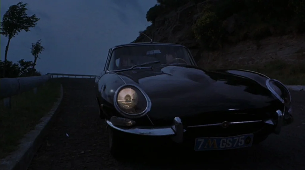 Danger Diabolik (1968) movie still: medium — A dark, winding road borders a steep rocky cliffside under a dim twilight sky. A black vintage sports…