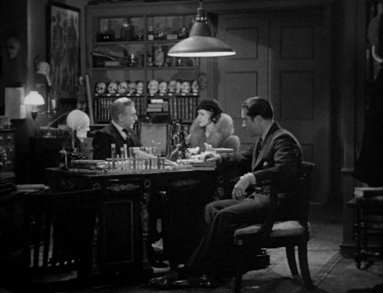 Frankenstein movie still: medium — A dark wood-paneled room is filled with medical curiosities including skulls and glassware. Three people sit at a…