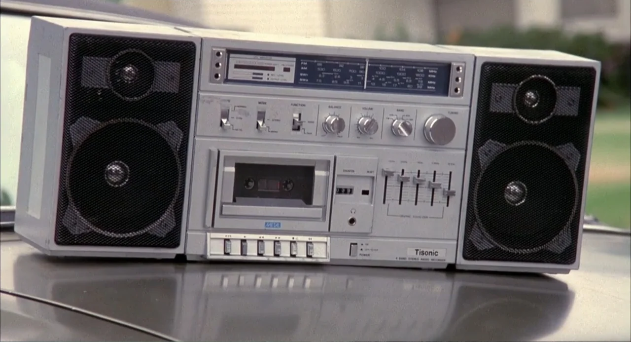 Teen Witch (1989) movie still: closeup — A silver Tisonic boombox is placed centrally on the shiny metallic hood of a car. The background shows…