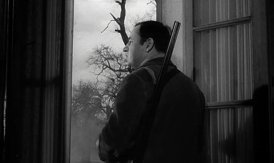 Thérèse Desqueyroux (1962) movie still: medium — A man stands inside a room looking out through a large, paned glass window. Beyond the glass, bare…