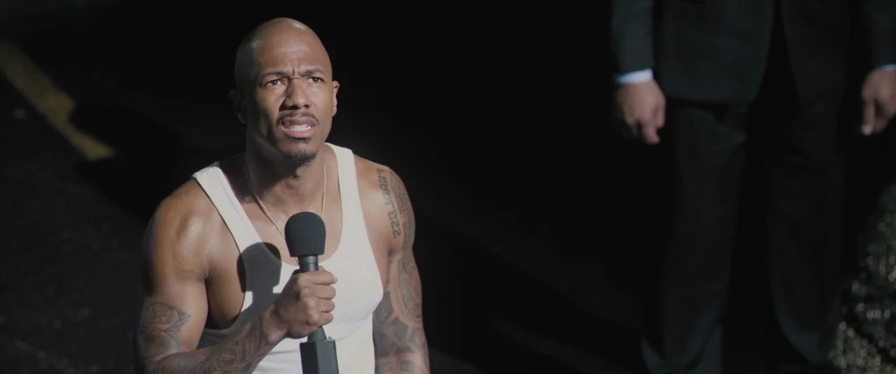 Chi-raq (2015) movie still: medium — A dark, possibly wet asphalt surface at night serves as the primary setting. A man in a tank top holds a…