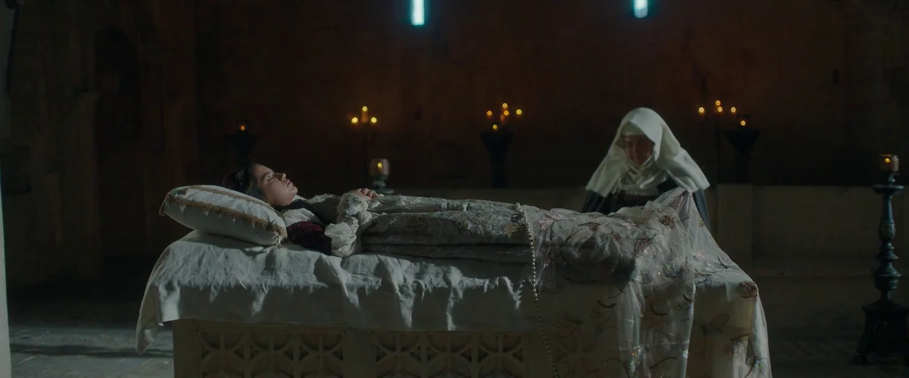 Rosaline (2022) movie still: wide — A stone chamber features a woman lying on a raised stone bier covered in linens. A nun kneels at the side of the…