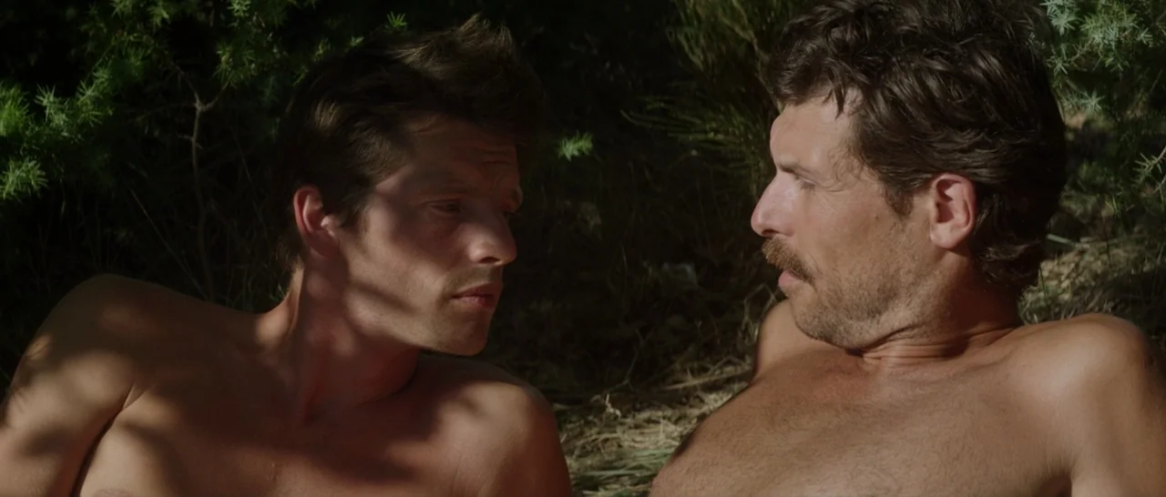 Stranger By The Lake (2013) movie still: medium — Two men lie shirtless on the shaded ground beneath dense tree foliage. They are face-to-face in a…