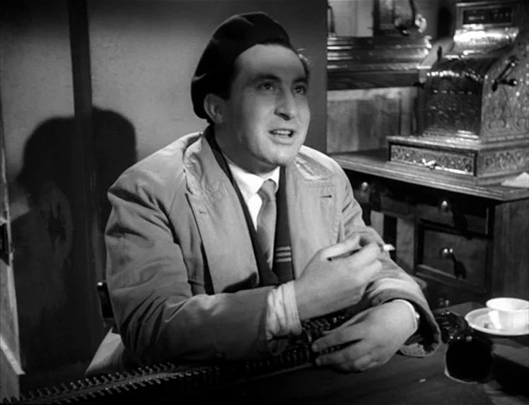 I Vitelloni (1953) movie still: medium — A man sits at a desk in a dark room with an old cash register. He is wearing a beret and a coat while…