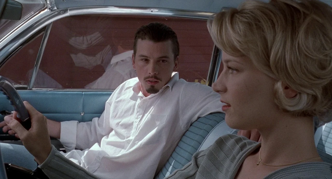Touch (1997) movie still: medium — The interior of a vintage car with blue upholstered seating. A man sits in the passenger seat observing a woman…