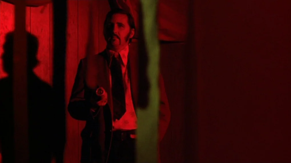 Mean Streets (1973) movie still: medium — A man stands in a dimly lit, narrow corridor featuring vertical wooden paneling. He holds a small,…