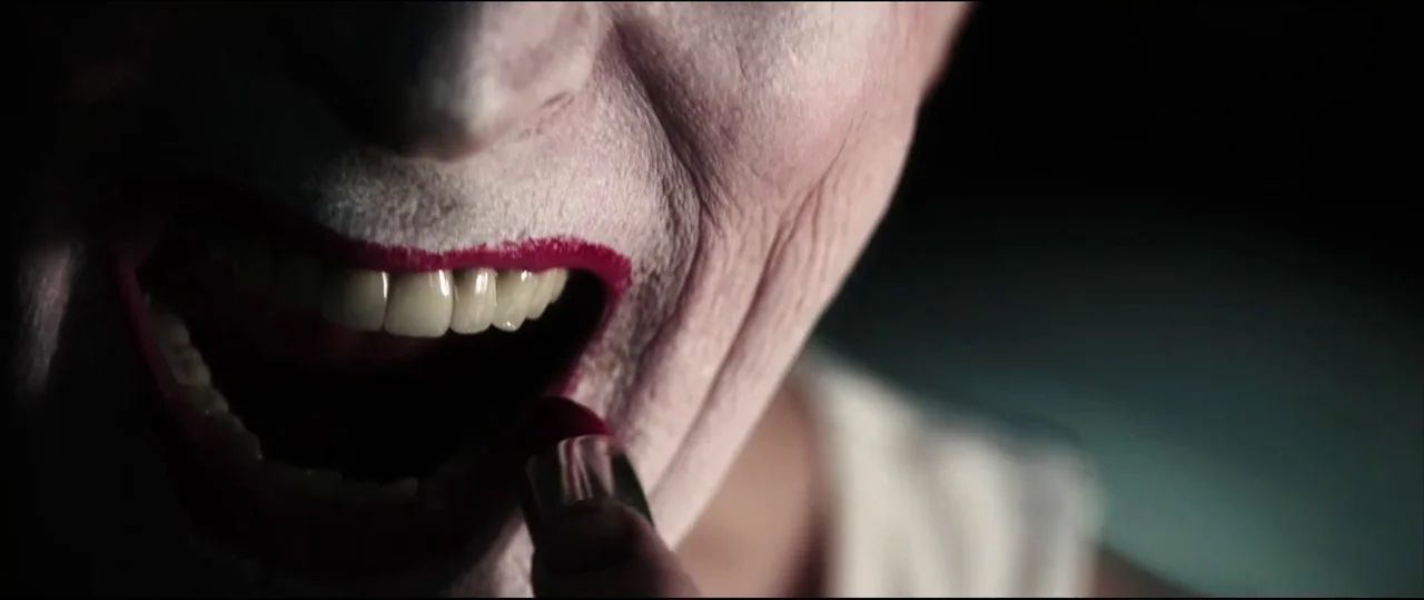 Insidious: Chapter 2 (2013) movie still: closeup — A close view of a person with white face makeup applying red lipstick. The lighting is…
