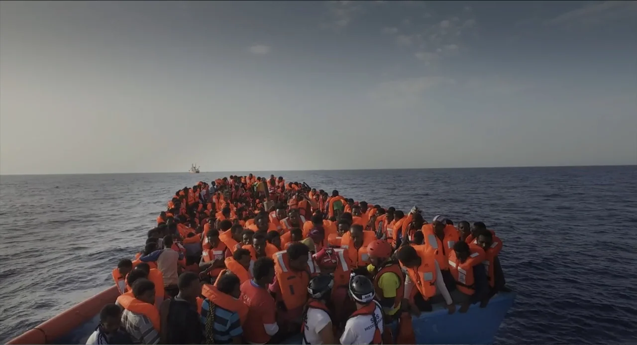 Human Flow (2017) movie still: wide — A large crowd of people sits packed tightly on a wooden boat at sea. Every person is wearing a bright orange…