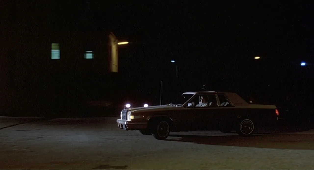 Death Wish II (1982) movie still: wide — An empty asphalt parking lot at night is illuminated by a car's bright headlights. A single sedan drives…