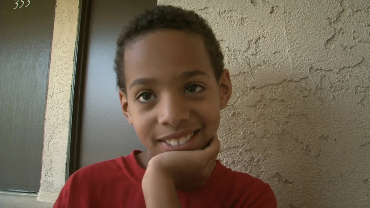 Homeless: The Motel Kids of Orange County (2010) movie still: closeup — A child stands outdoors in front of a tan stucco wall next to a brown door.…