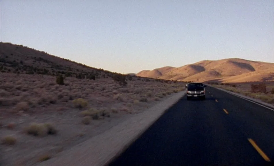 The Brown Bunny (2003) movie still: wide — An asphalt road cuts through a vast, arid desert landscape with rolling brown hills. A dark minivan…