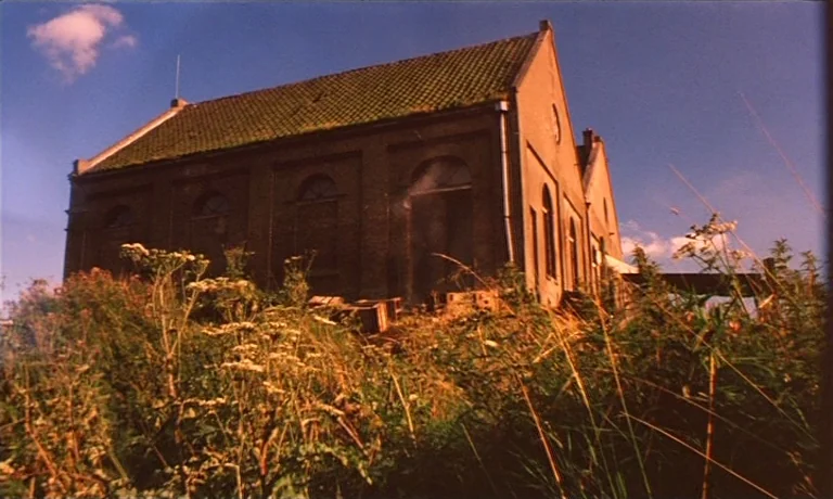 Katie Tippel (1975) movie still: establishing — An aged brick building with a mossy roof stands amidst overgrown tall grasses. The scene is…