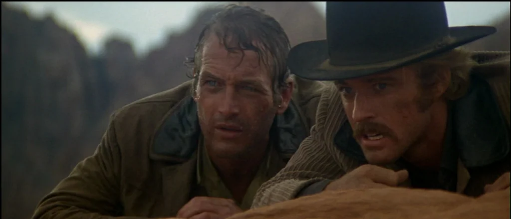 Butch Cassidy and the Sundance Kid (1969) movie still: medium — Two men hide behind a desert ridge composed of eroded sandstone and rock. Both men…