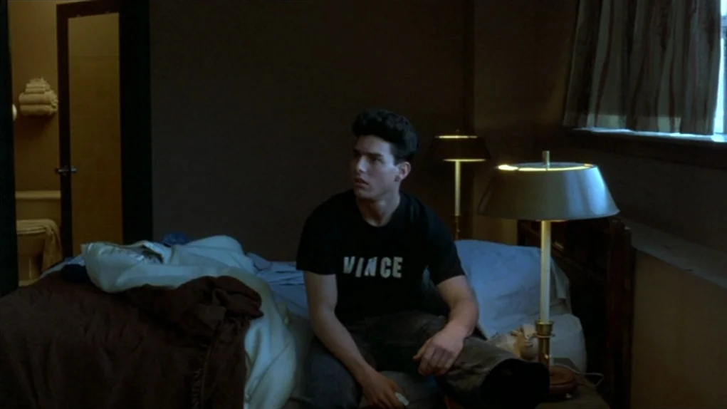 The Colour of Money (1986) movie still: medium — A dim, dated motel room features a bed and a visible bathroom entrance. A young man sits on the bed…