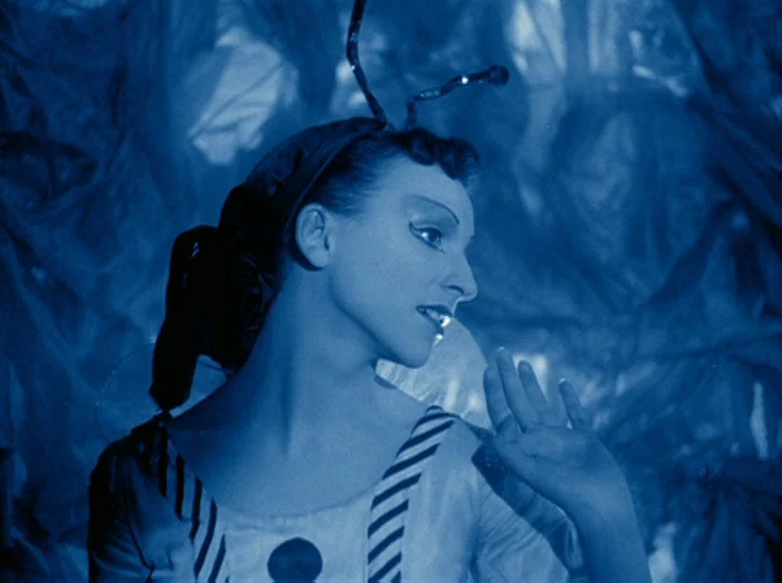 Rabbit’s Moon (1950) movie still: medium — A woman wears an insect costume with antennae against a crinkled, translucent background. She raises her…