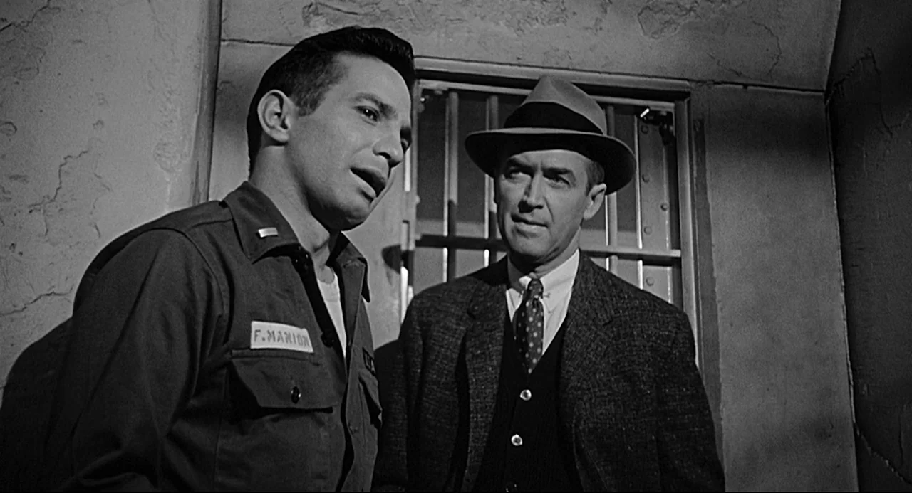 Anatomy of a Murder (1959) movie still: medium — A dimly lit room with stone walls and a metal grate in the background. Two men stand facing each…