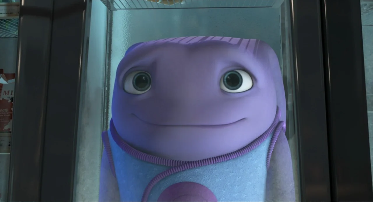 Home (2015) movie still: closeup — A purple, rounded, alien creature is positioned inside a brightly lit refrigerator. The character stares directly…