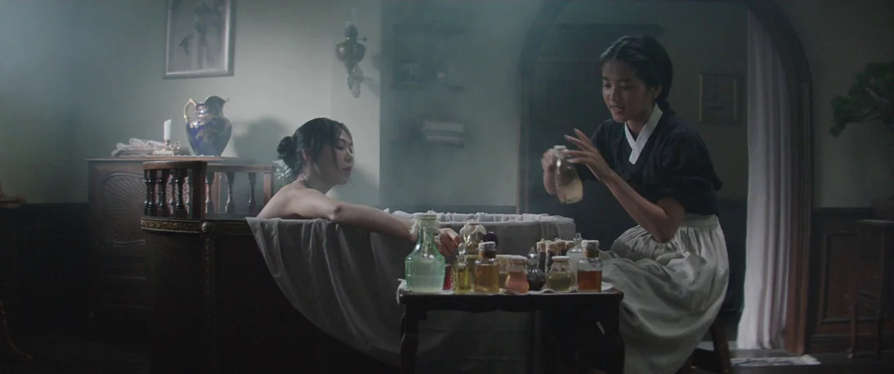 The Handmaiden (2016) movie still: medium — A dimly lit room contains a wooden bathtub and a small table with bottles. A woman in a black top pours…