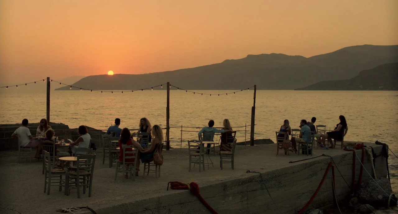 Before Midnight (2013) movie still: wide — A concrete pier extends into calm water under a deep orange sunset sky. Small tables and chairs are…
