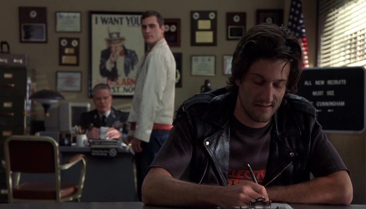 Signs (2002) movie still: medium — A military recruitment office wall is covered in framed certificates and a propaganda poster. A young man in a…