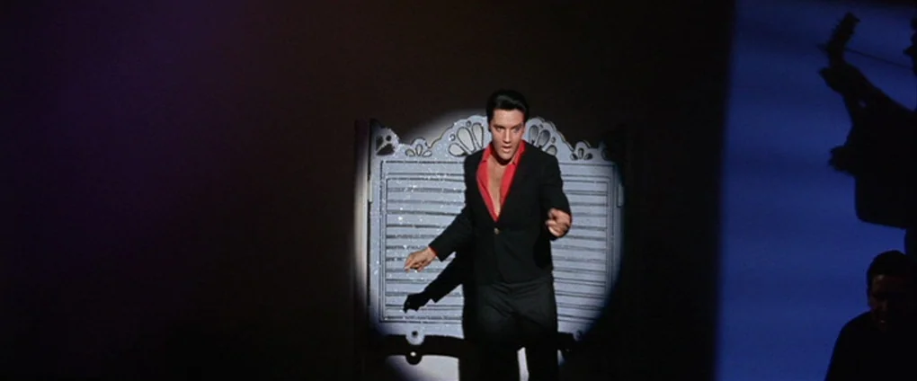 Viva Las Vegas (1964) movie still: fullbody — A dark stage features a single white spotlight on a man in black. He stands before a stylized lattice…