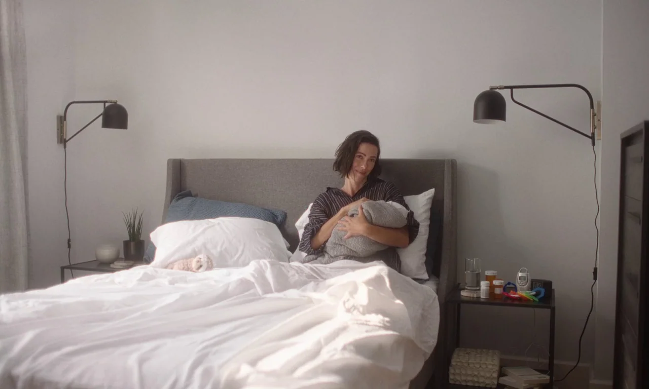 Resurrection (2022) movie still: medium — A light-walled room contains a grey upholstered bed and twin dark metal wall lamps. A woman sits propped…