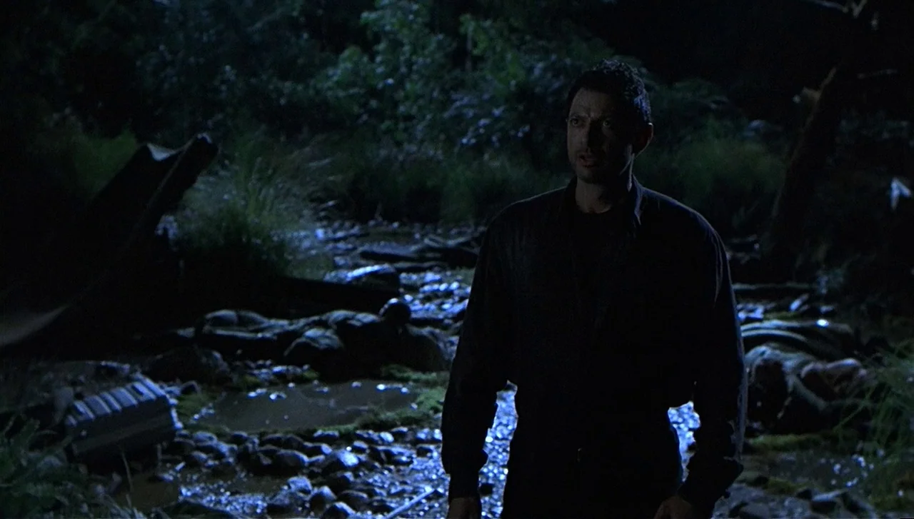 The Lost World: Jurassic Park (1997) movie still: medium — A dark, wooded stream bed at night with dense foliage and wet rocks. A man in a dark…