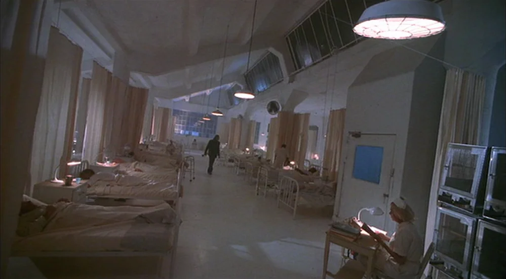 The Fisher King (1991) movie still: wide — A clinical hospital ward contains multiple rows of beds separated by thin, hanging curtains. A lone nurse…